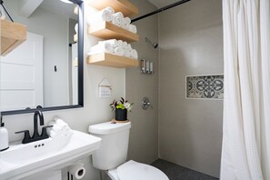 A modern bathroom with a white sink, black faucet, and a mirror above it. There are wooden shelves holding rolled towels, a toilet, and a shower with a glass door. - A pet friendly A-Frame with a hot tub & fire pit! (McAllen)