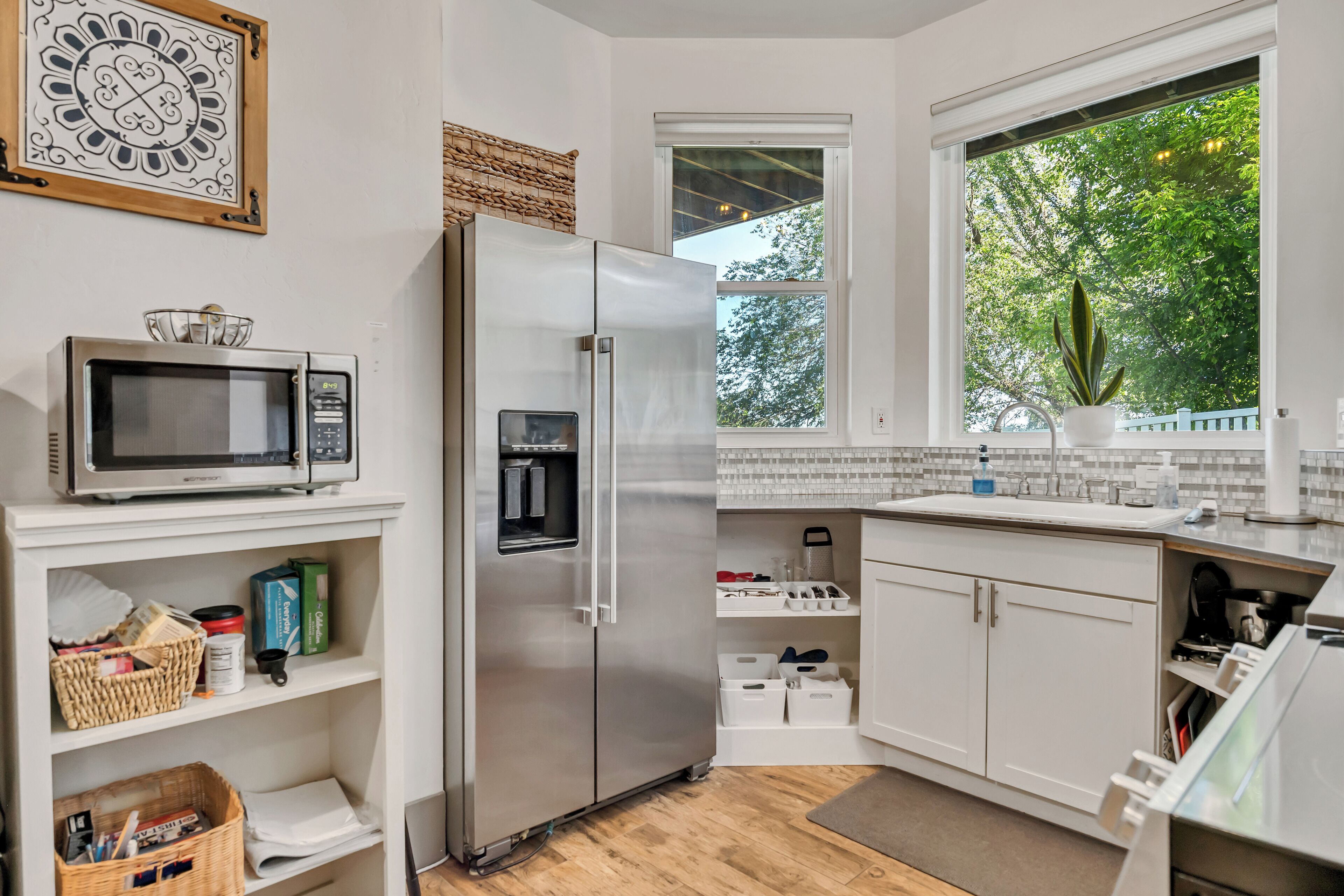 A modern kitchen with white cabinetry, a stainless steel refrigerator, a microwave, and a window with a view of trees.