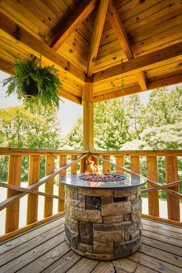 A covered outdoor deck with a stone fire pit, wooden railings, and hanging plants.