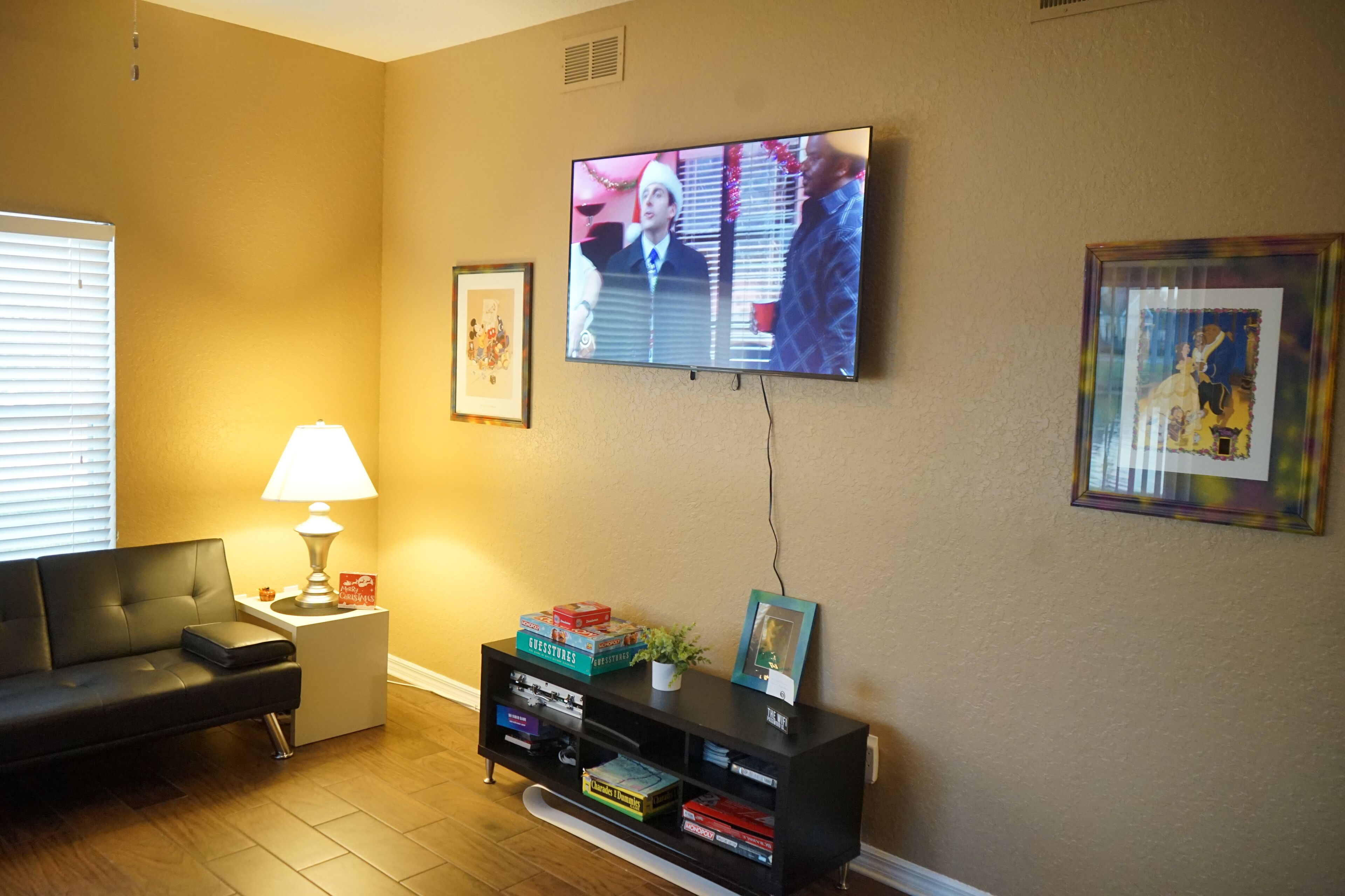A living room with a flat-screen TV mounted on the wall, a black sofa, a side table with a lamp, and a bookshelf with various items.