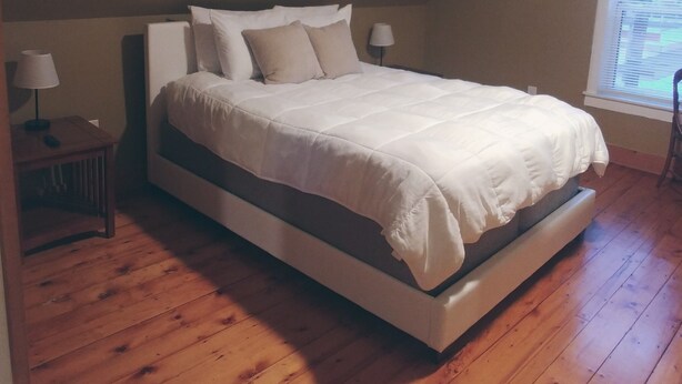 Een slaapkamer met een groot bed, houten vloeren, een nachtkastje met een lamp en een raam met luxaflex.
