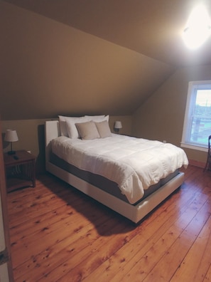 A bedroom with a large bed, wooden flooring, a nightstand with a lamp, and a window with blinds.