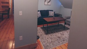 Een kamer met een houten vloer, een zwarte bank met gestreepte kussens, een klein houten tafeltje en een ingelijst kunstwerk aan de muur.