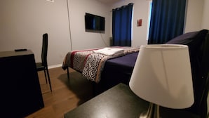Una habitación pequeña y ordenada con cama, escritorio y silla. Hay un televisor montado en la pared y una ventana con cortinas.