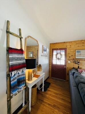 A cozy living room with a wooden floor, a sofa, a lamp, a mirror, and a door with a window.