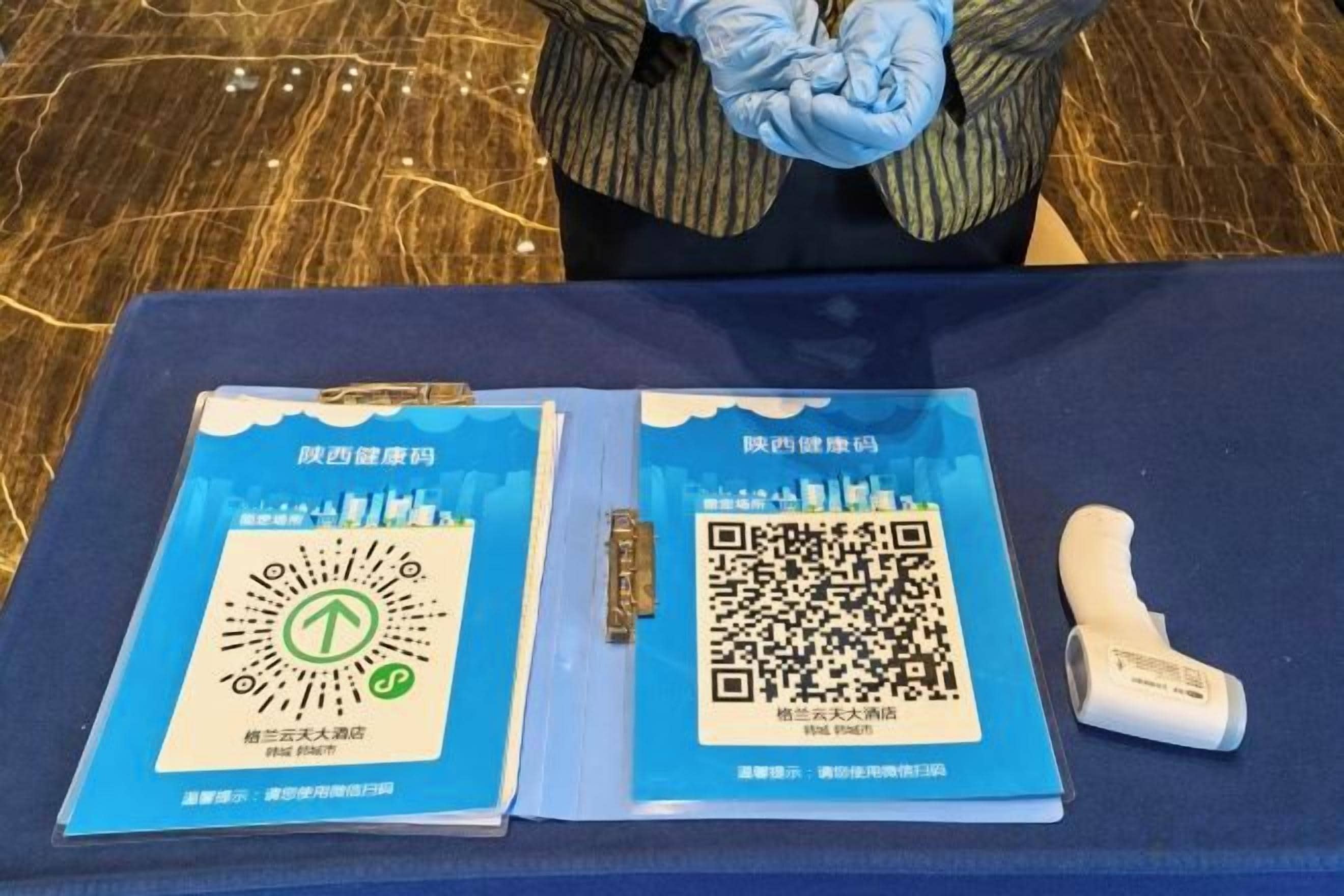 Une table avec deux dossiers bleus contenant des codes QR et du texte, un objet blanc ressemblant à une télécommande, et une personne à l’arrière-plan.