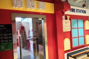 Entrada de una estación de bomberos con un letrero amarillo, un reloj digital que marca 88:88, y un interior colorido con un banco y un tren de juguete.