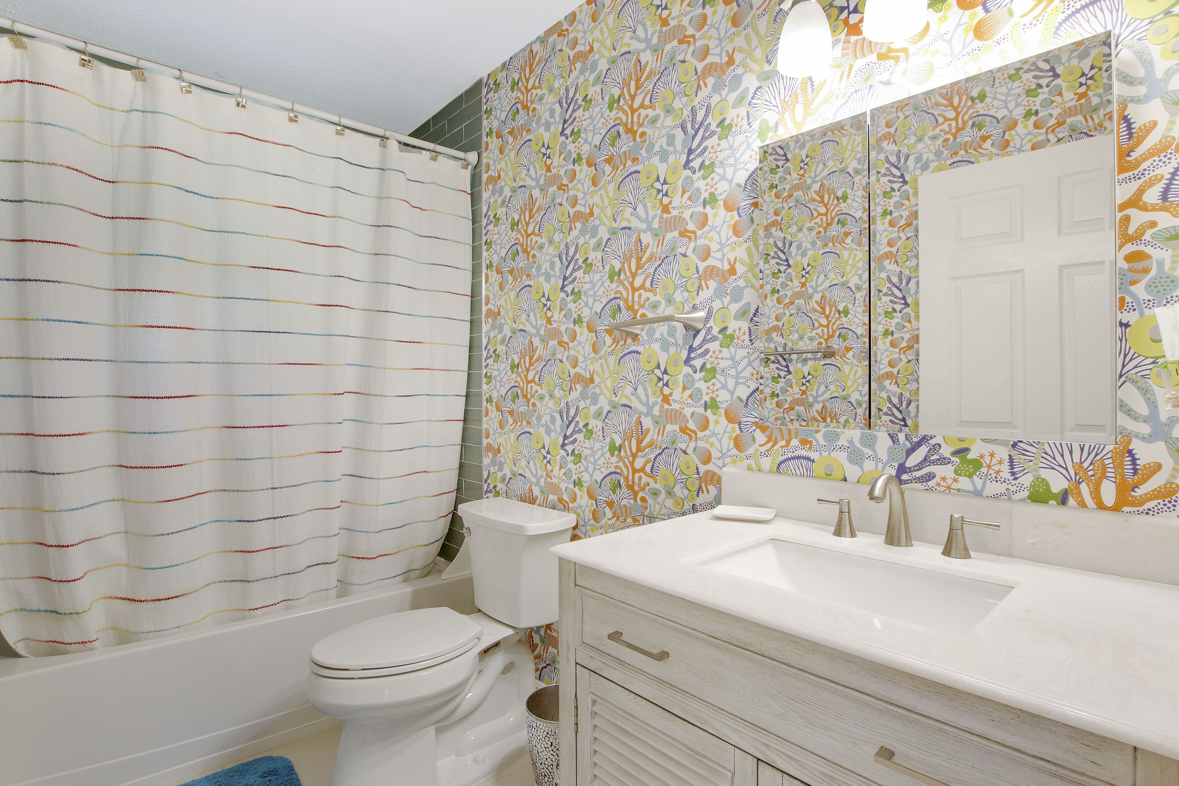 A bathroom with a striped shower curtain, a toilet, a sink with a mirror, and a tiled wall with a coral pattern.