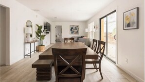 Un vaste espace dédié à la salle à manger, avec une longue table en bois et des chaises, une petite table d’appoint avec une lampe, et une grande fenêtre offrant une vue sur l’extérieur.