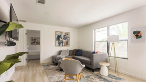 Un salon moderne avec un canapé gris, une petite table ronde et un lampadaire. Un tableau est accroché au mur, et une grande fenêtre est dotée de stores.