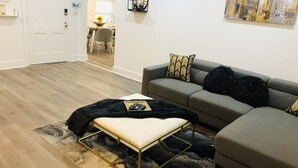 Un salon moderne avec un canapé d’angle gris, un pouf noir et blanc, et un tableau représentant un paysage urbain accroché au mur.
