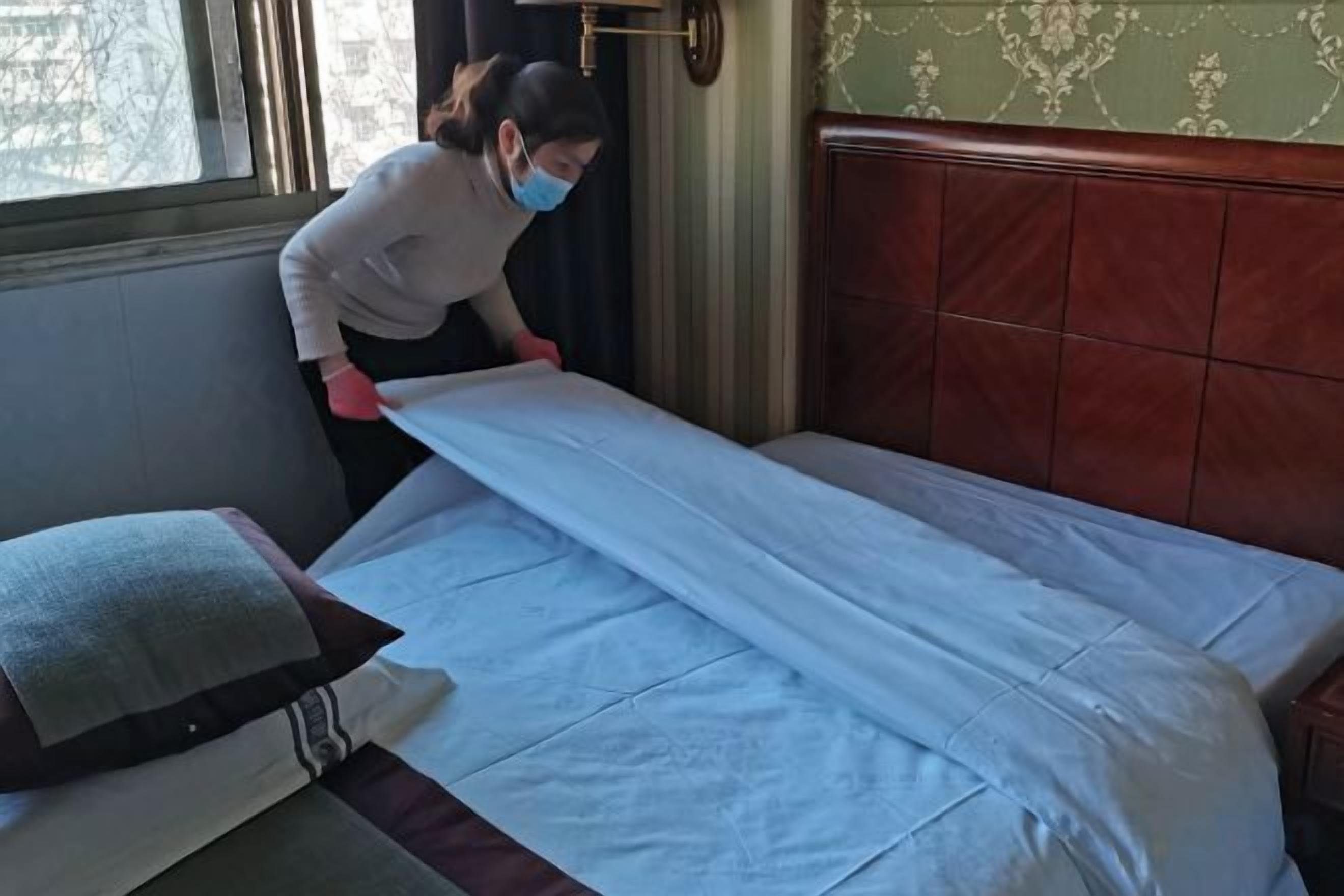 A person adjusting a bedspread in a room with a wooden headboard and a window.