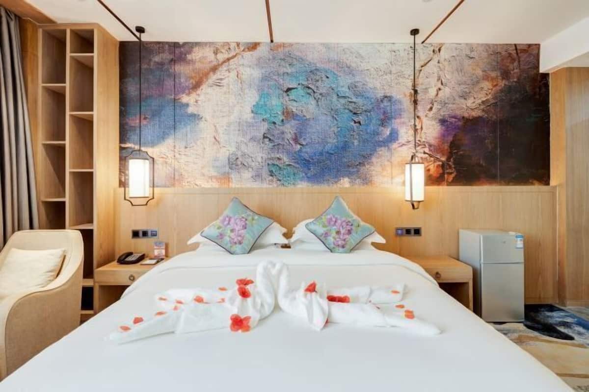 A hotel room with a large abstract painting on the wall, a bed with floral pillows, a bedside table with a lamp, and a small refrigerator.