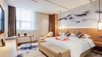 A modern hotel room with a large bed, a desk, a chair, and a bookshelf.