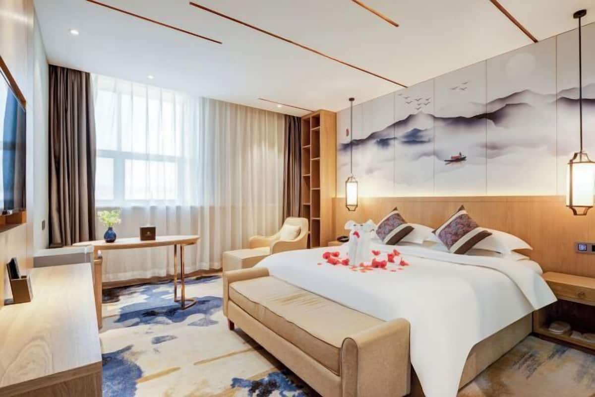 A modern hotel room with a large bed, a desk, a chair, and a bookshelf.