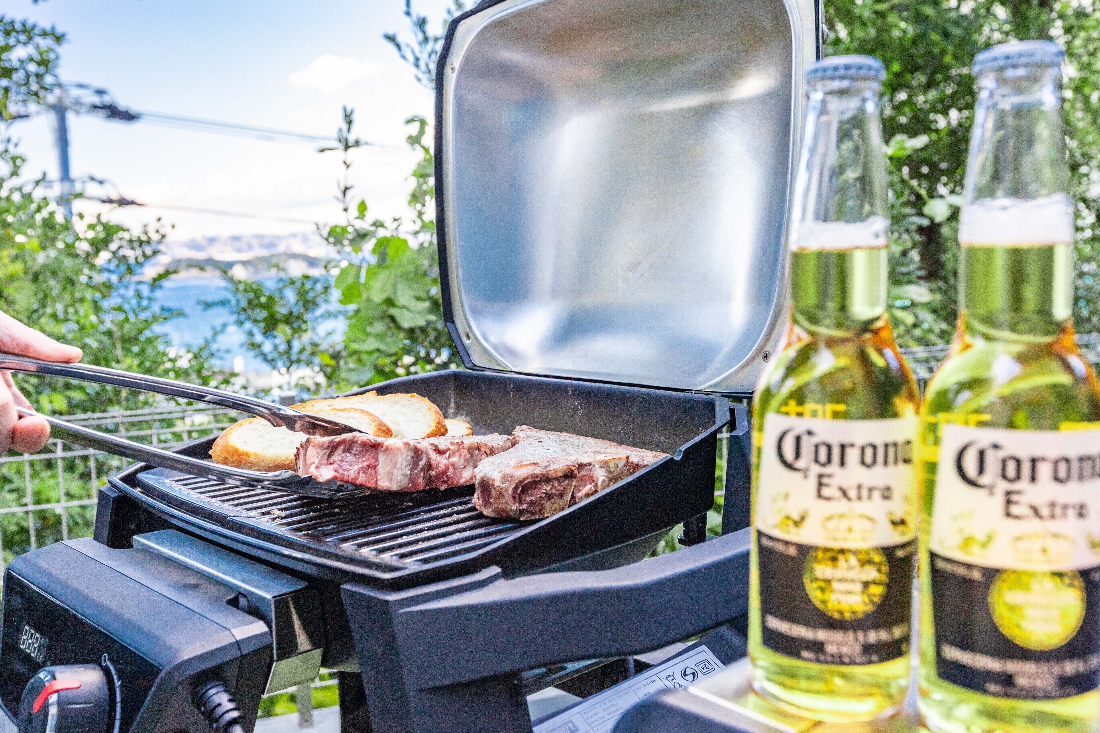 Uma churrasqueira com bife e pão, e duas garrafas de cerveja Corona Extra.
