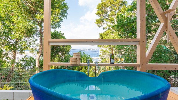 Uma banheira de hidromassagem azul em um deck de madeira, com vista para árvores e um corpo d'água.