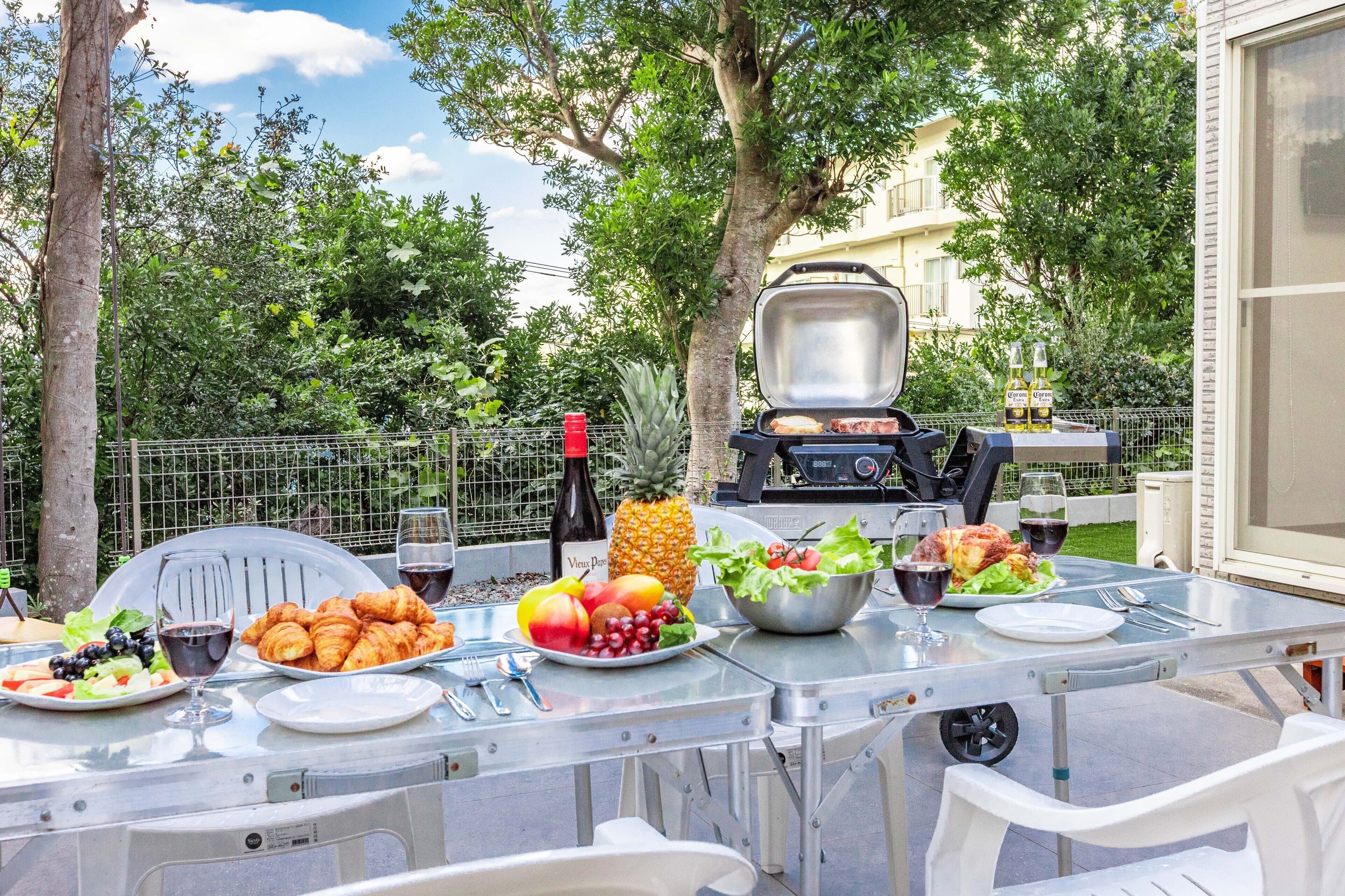 Un área de comedor al aire libre con una parrilla, una mesa dispuesta con comida y bebidas, y una vista de un edificio al fondo.
