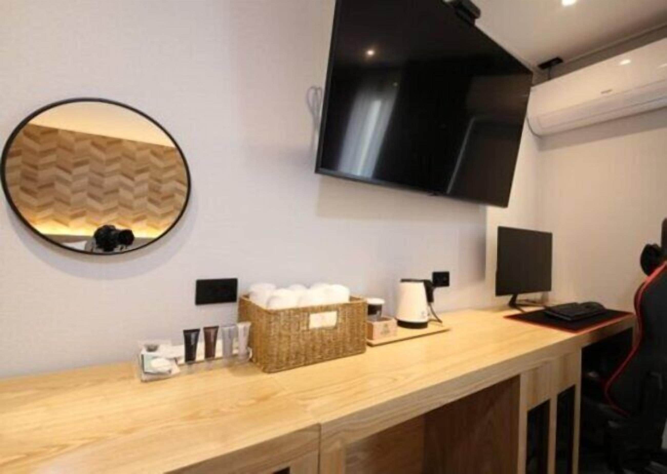 Una habitación de hotel moderna con un escritorio de madera, un espejo redondo, un televisor de pantalla plana montado en la pared, una mesita con una tetera y una cesta de artículos de aseo.
