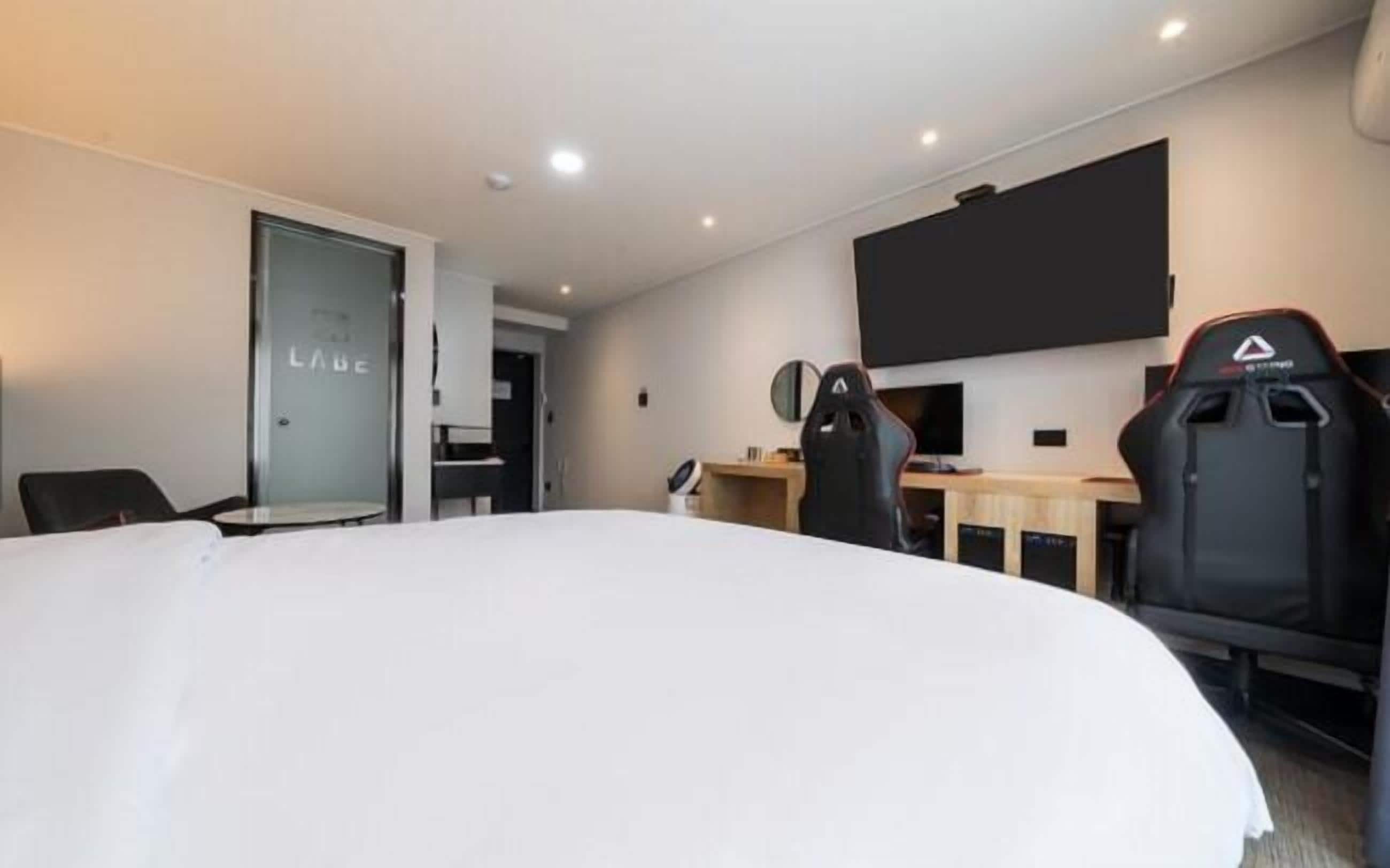 A hotel room with a large bed, a desk with a computer, two gaming chairs, and a flat-screen TV mounted on the wall.