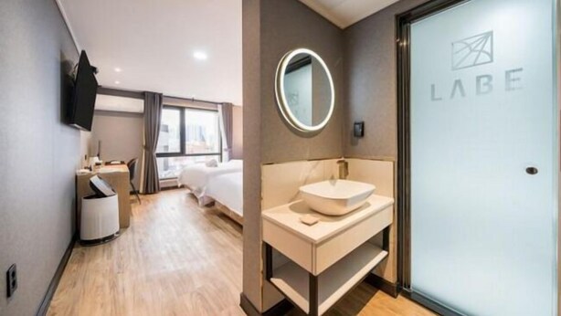 A hotel room with a bed, a bedside table, a mirror, a bathroom with a sink, and a frosted door with the logo "LABE". - LABE Hotel (Jeonju)