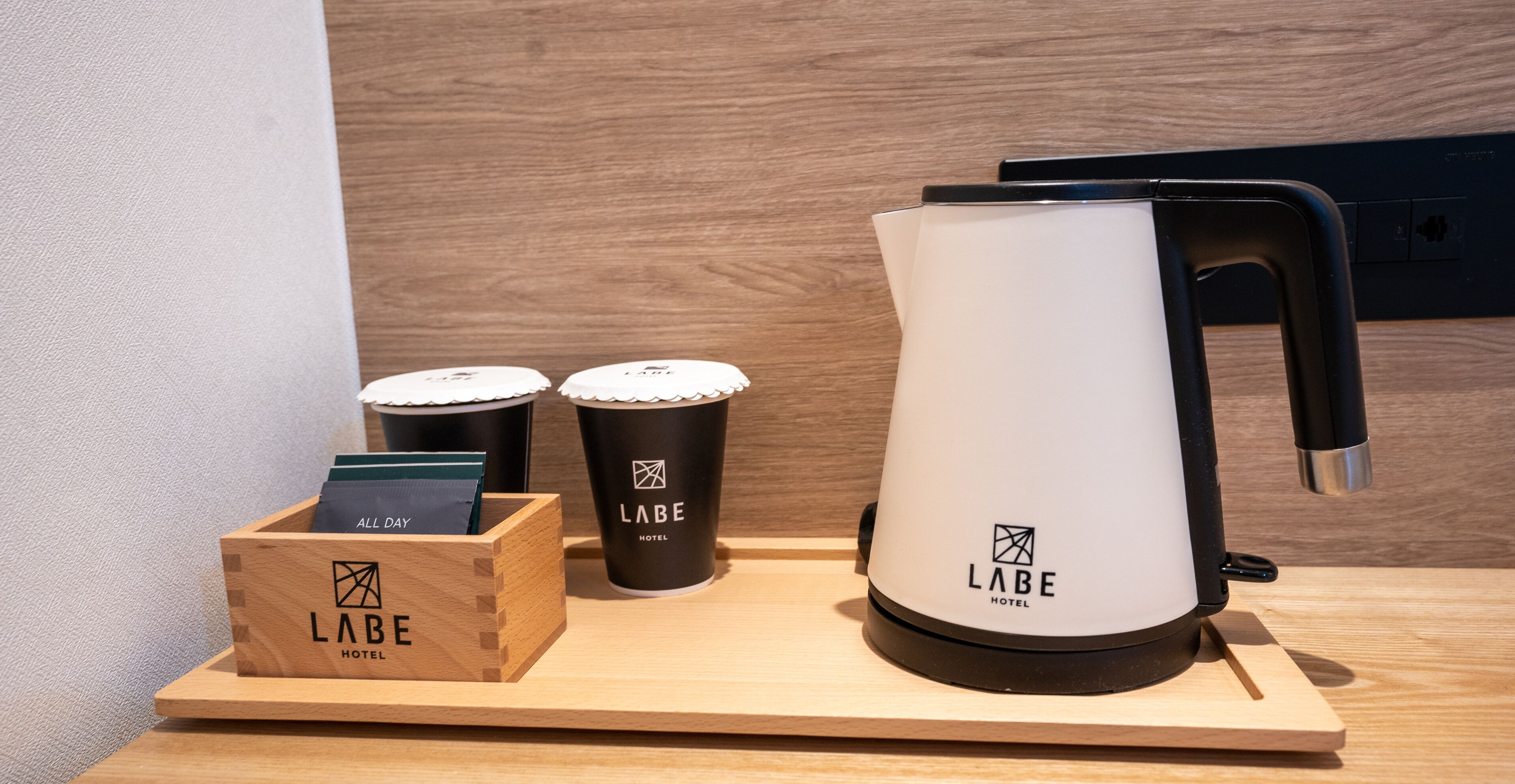 Una estación de café con cafetera, dos vasos desechables y una caja de madera con una tarjeta y una identificación.