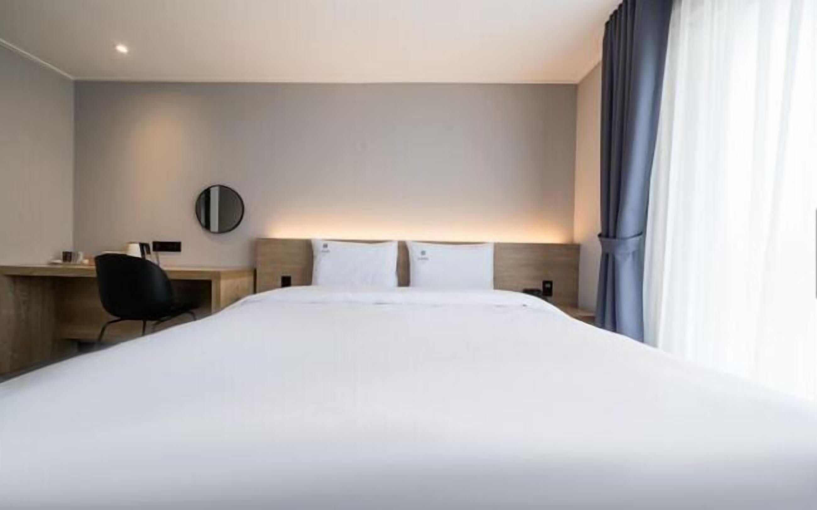 Habitación de hotel con una cama grande, un escritorio con silla, un espejo redondo y una ventana con cortinas.