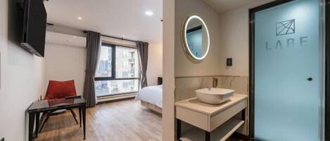 A modern hotel room with a bed, a desk, a round mirror, a sink, and a door with the logo "LABE".