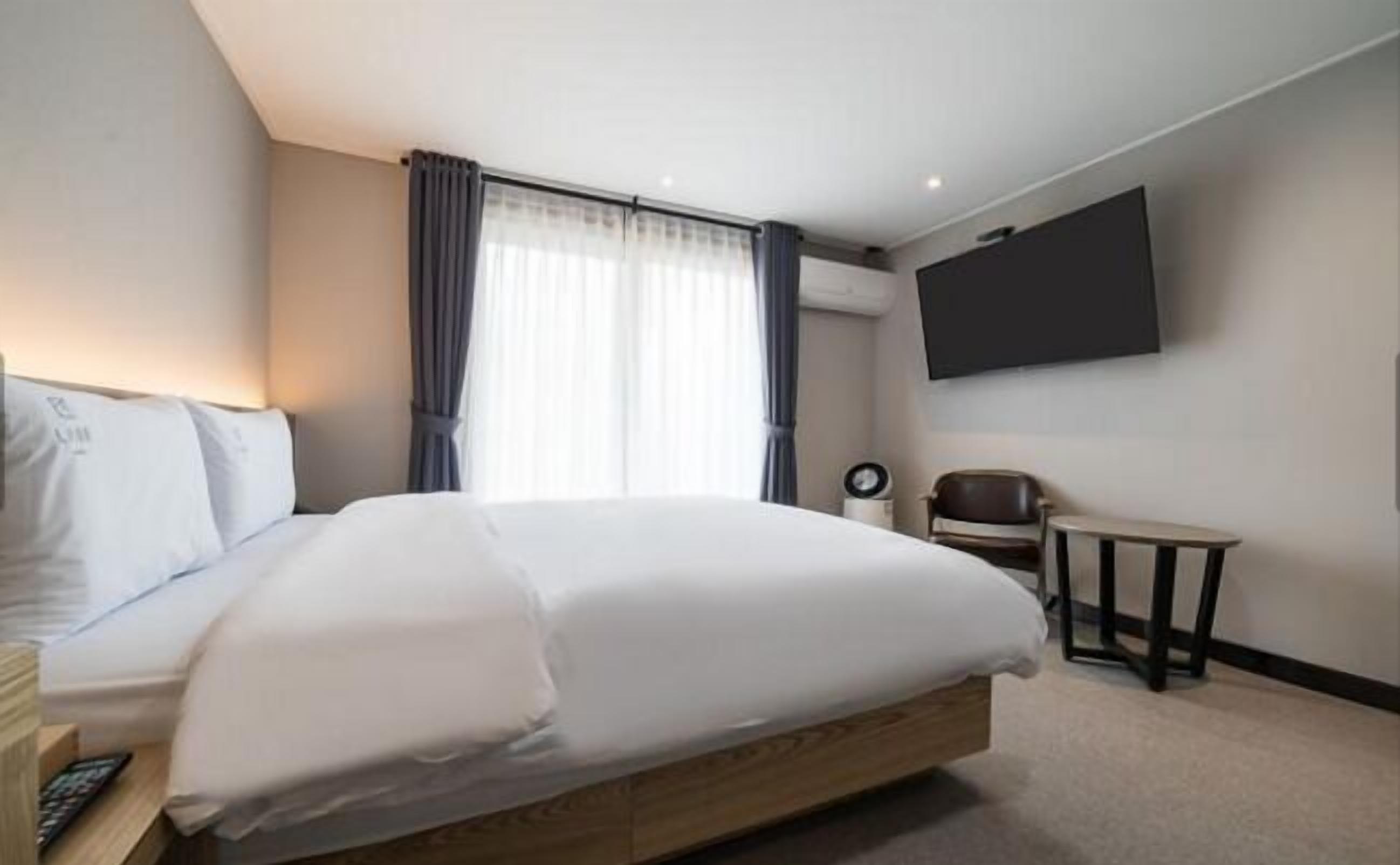 Quarto de hotel com uma cama grande, televisão na parede, uma mesinha e uma cadeira.