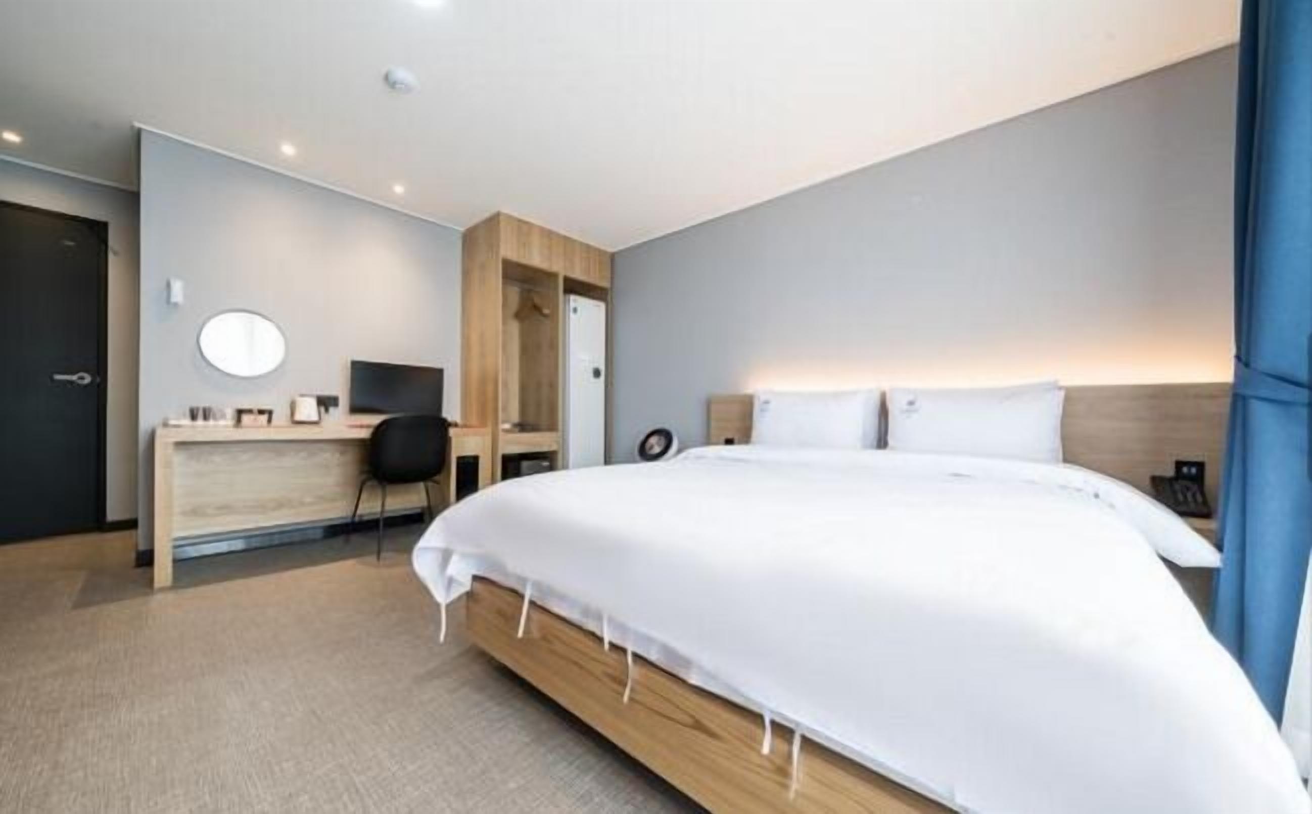 A modern hotel room with a large bed, a desk with a chair, a round mirror, and a wardrobe.