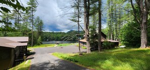 Una cabaña con un camino de entrada pavimentado que lleva a un muelle junto a un lago, rodeada de un bosque frondoso y una montaña al fondo.