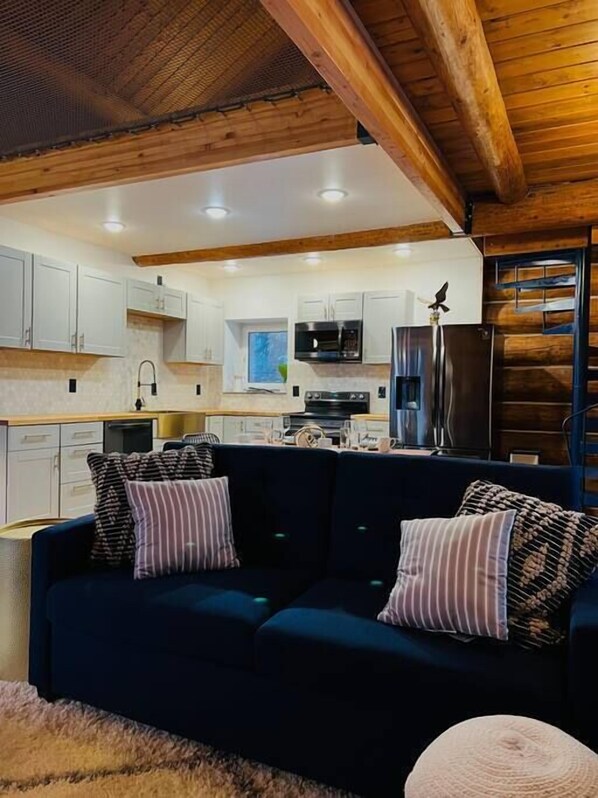 A cozy living area with a dark blue sofa, striped pillows, and a round ottoman. A wooden ceiling and white cabinets are visible in the background. - This beautiful log cottage has large windows that let in lots of light (Fairbanks)