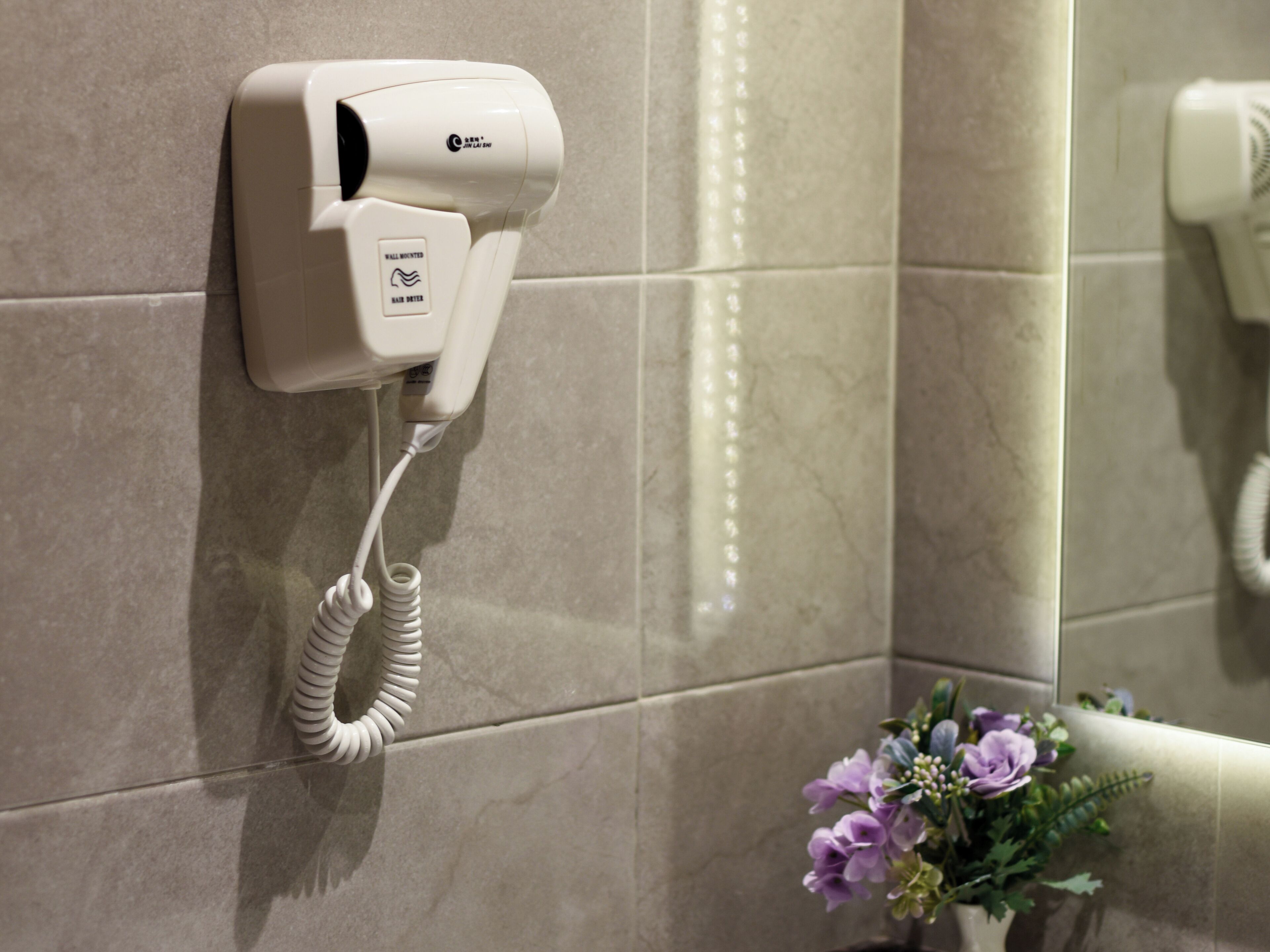 A wall-mounted hair dryer in a bathroom with a tiled wall and a flower arrangement in the corner.