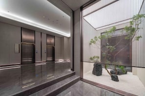 A modern elevator area with three elevators, a polished marble floor, and a view of a landscaped garden through a glass partition.