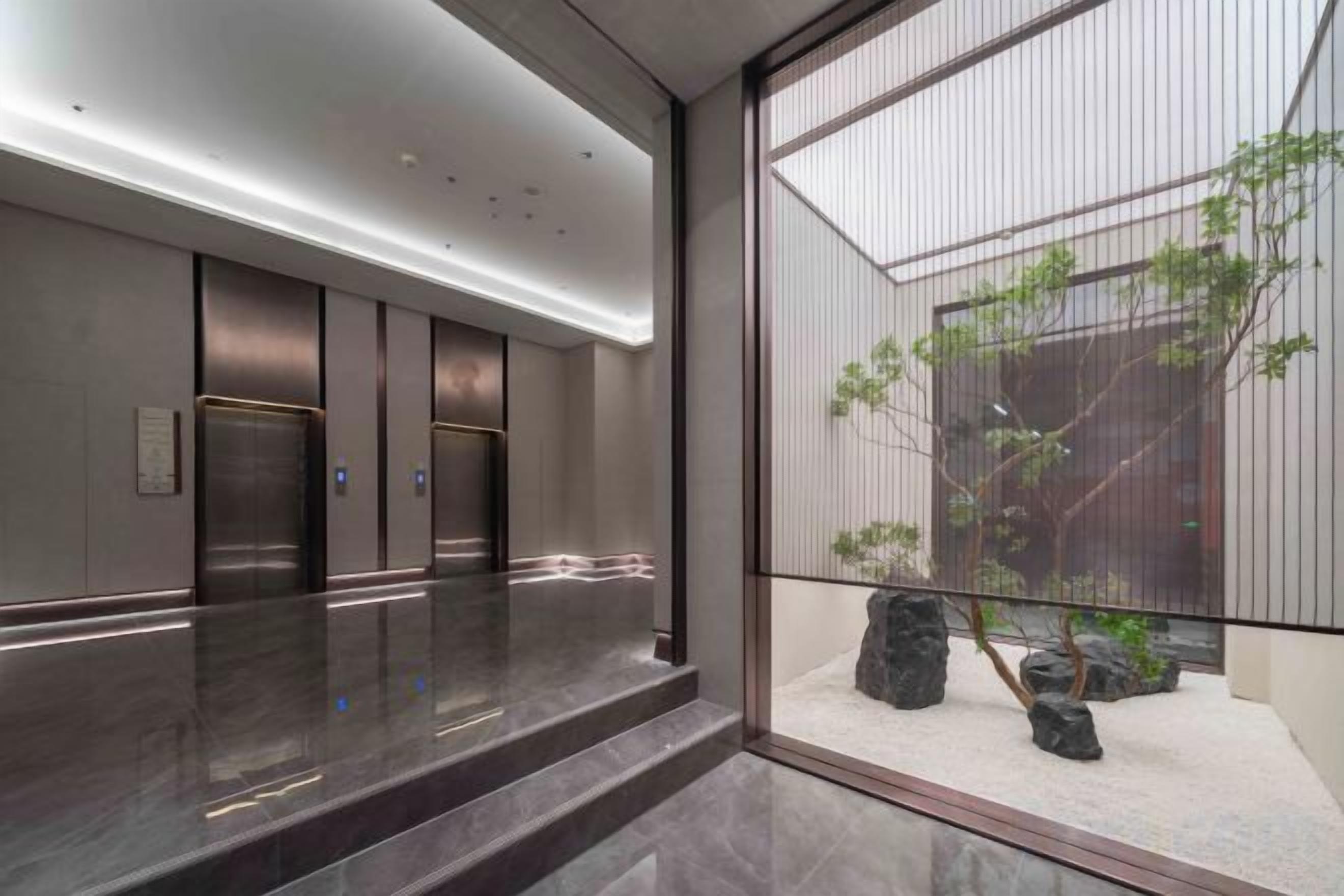 A modern elevator area with a reflective floor, a glass partition, and a view of a landscaped outdoor area with rocks and plants.