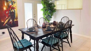 Un comedor con una mesa de madera oscura, seis sillas con cojines azules, un ventilador de techo y un cuadro grande en la pared.