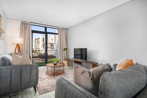 A modern living room with a sofa set, a coffee table, a TV, and a view of buildings through large windows. - Eden On the Bay 231 (Cape Town)