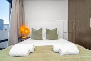 A hotel room with a bed, two pillows, a nightstand with a lamp, and a view of the outside. - Eden On the Bay 231 (Cape Town)