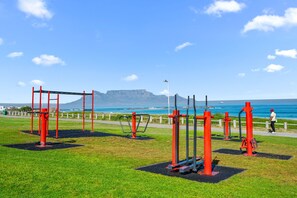 An outdoor gym with red exercise equipment, a paved path, and a scenic view of the ocean and a mountain in the background.