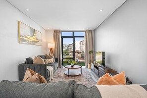 A modern living room with a grey sofa, a coffee table, and a TV. There is a large window with curtains, and a painting on the wall. - Eden On the Bay 231 (Cape Town)
