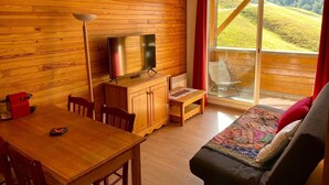 A cozy room with a wooden interior, a large window with a view of green hills, a flat-screen TV, a dining table with chairs, and a sofa with a colorful blanket. - 2 Rooms 35m2 South East view very well equipped (Allos)