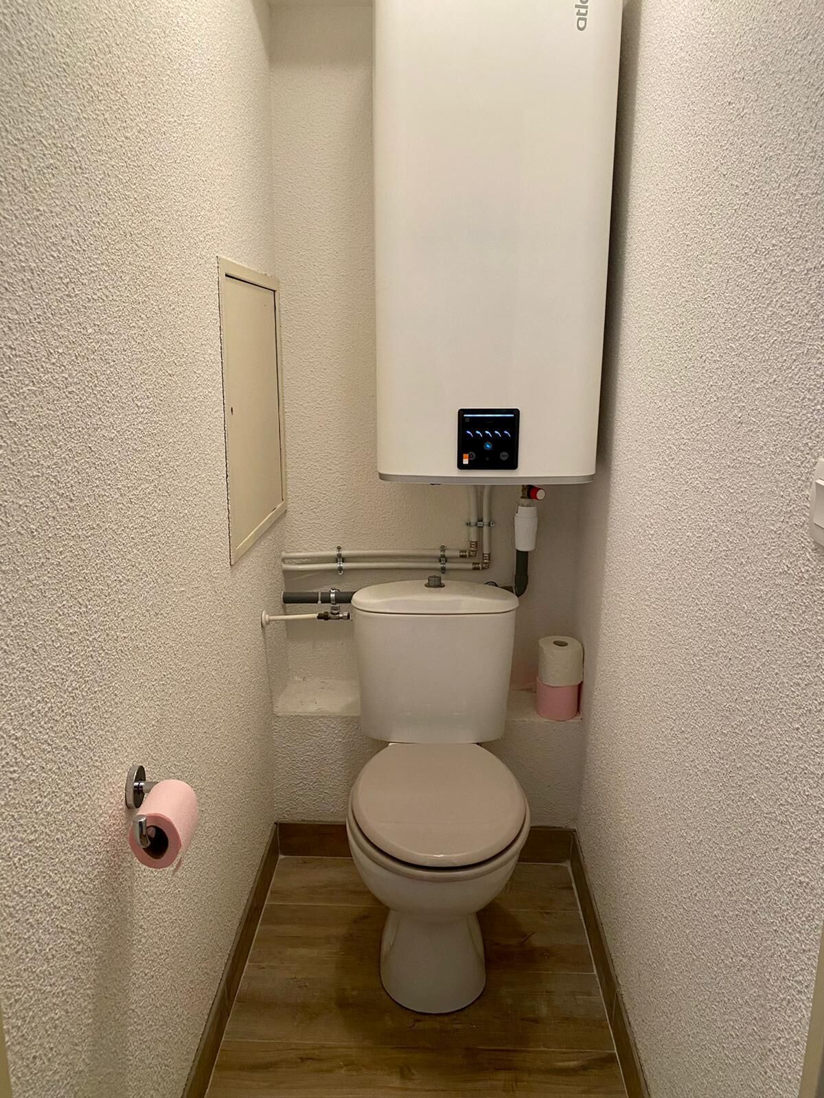 A small bathroom with a toilet, a white tank, a roll of toilet paper, and a pink toilet paper holder.