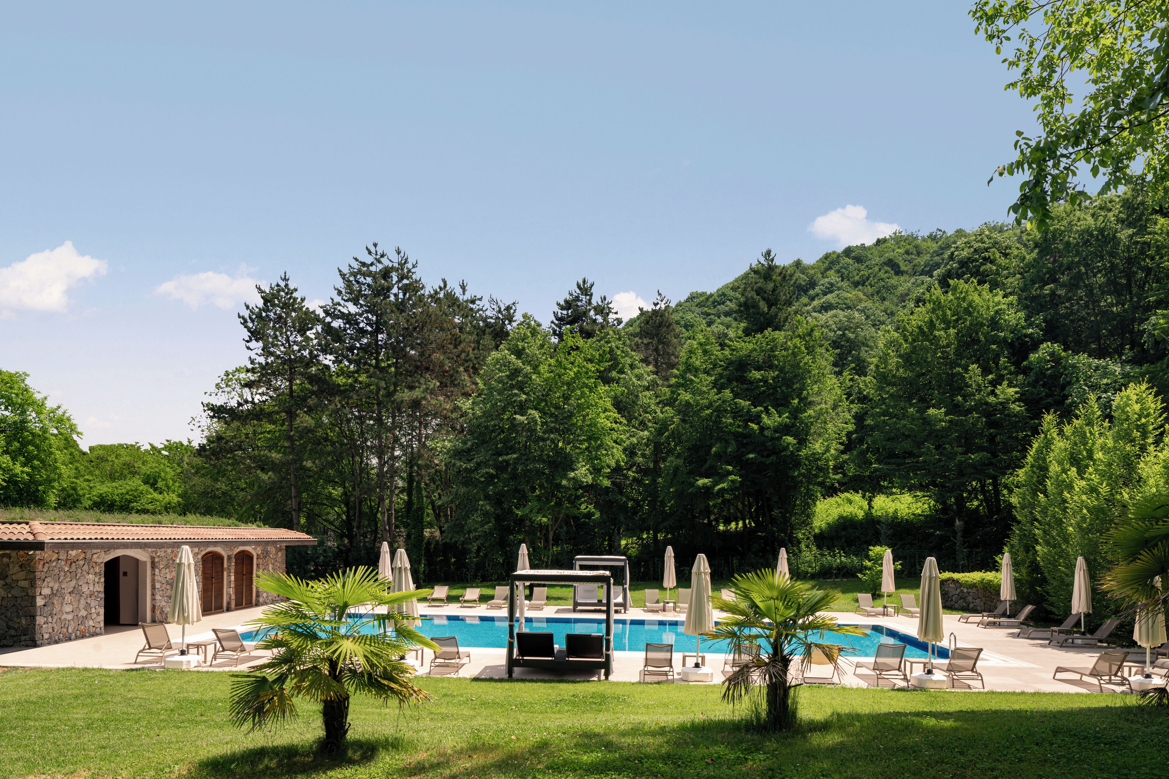 A pool area with lounge chairs and umbrellas, surrounded by lush greenery and a stone building.