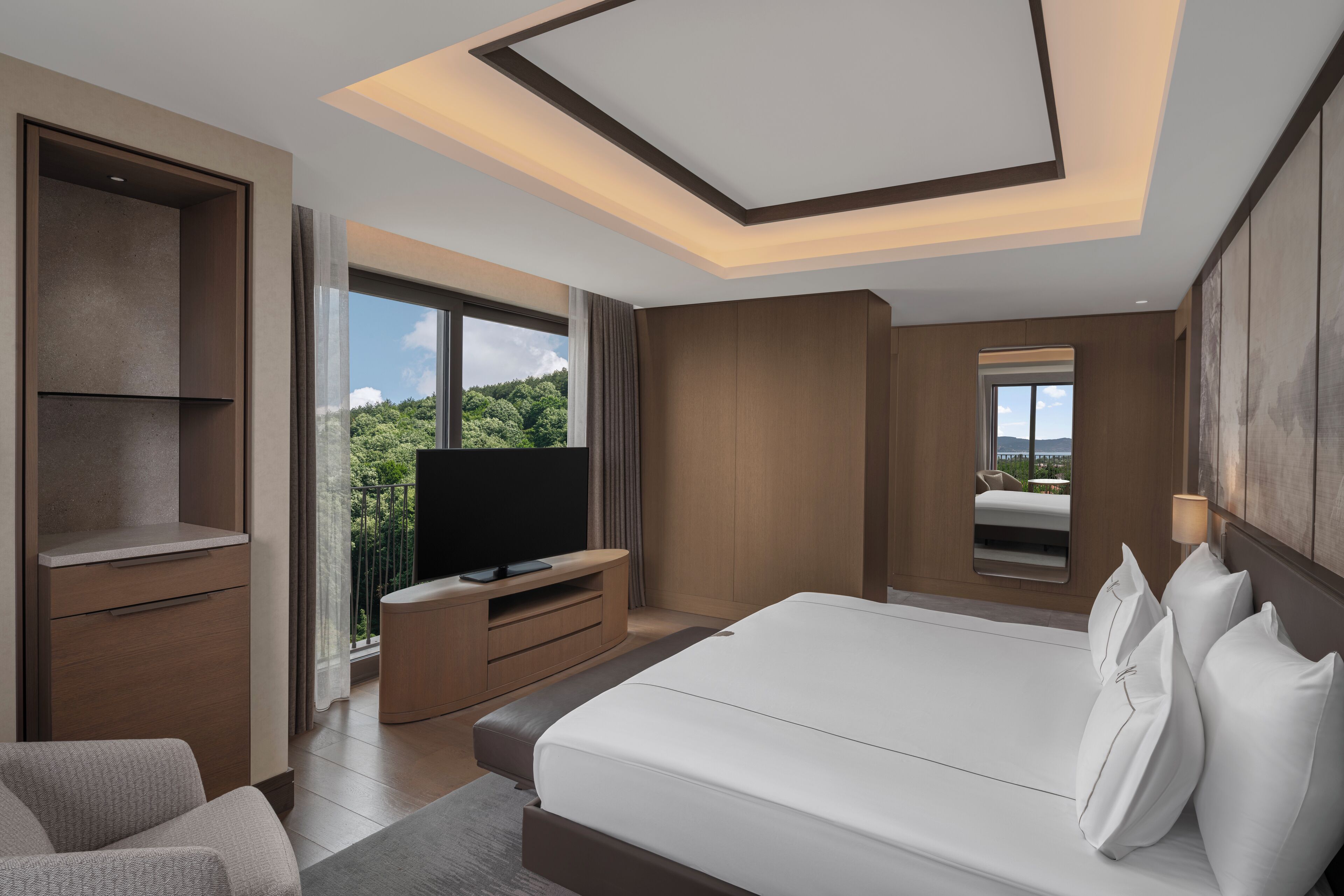 A modern hotel room with a large bed, a TV, a balcony with a view of greenery, and a wooden wardrobe.