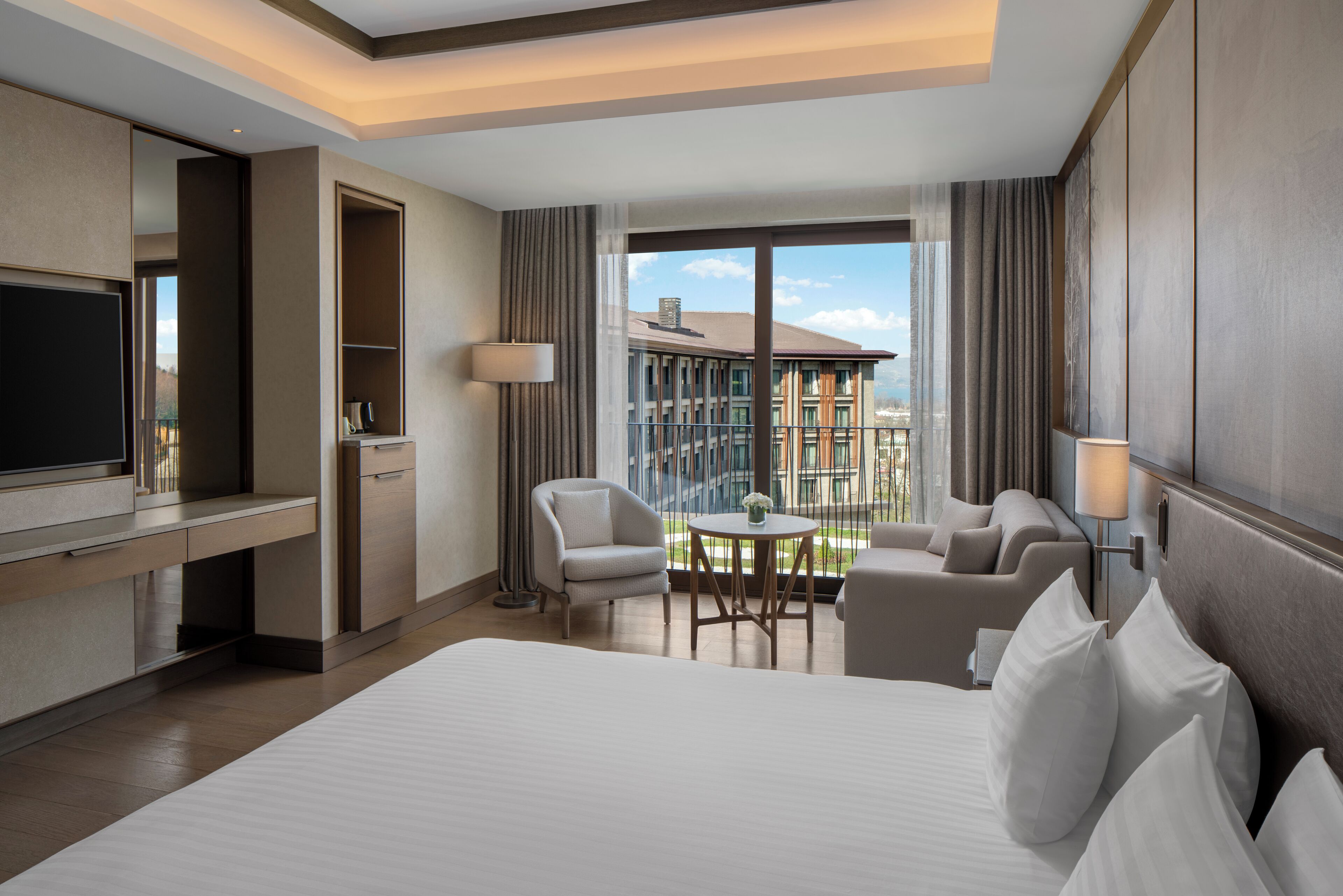 A hotel room with a large bed, a desk with a flat-screen TV, a seating area with a table and chairs, and a balcony with a view of buildings.