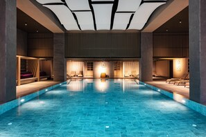A large indoor swimming pool with a modern design, featuring a central clock, lounge areas, and a ceiling with recessed lighting.