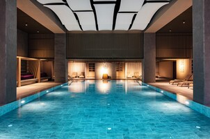 Une grande piscine intérieure au design moderne, dotée d’une horloge centrale, d’espaces de détente et d’un plafond avec des luminaires encastrés.