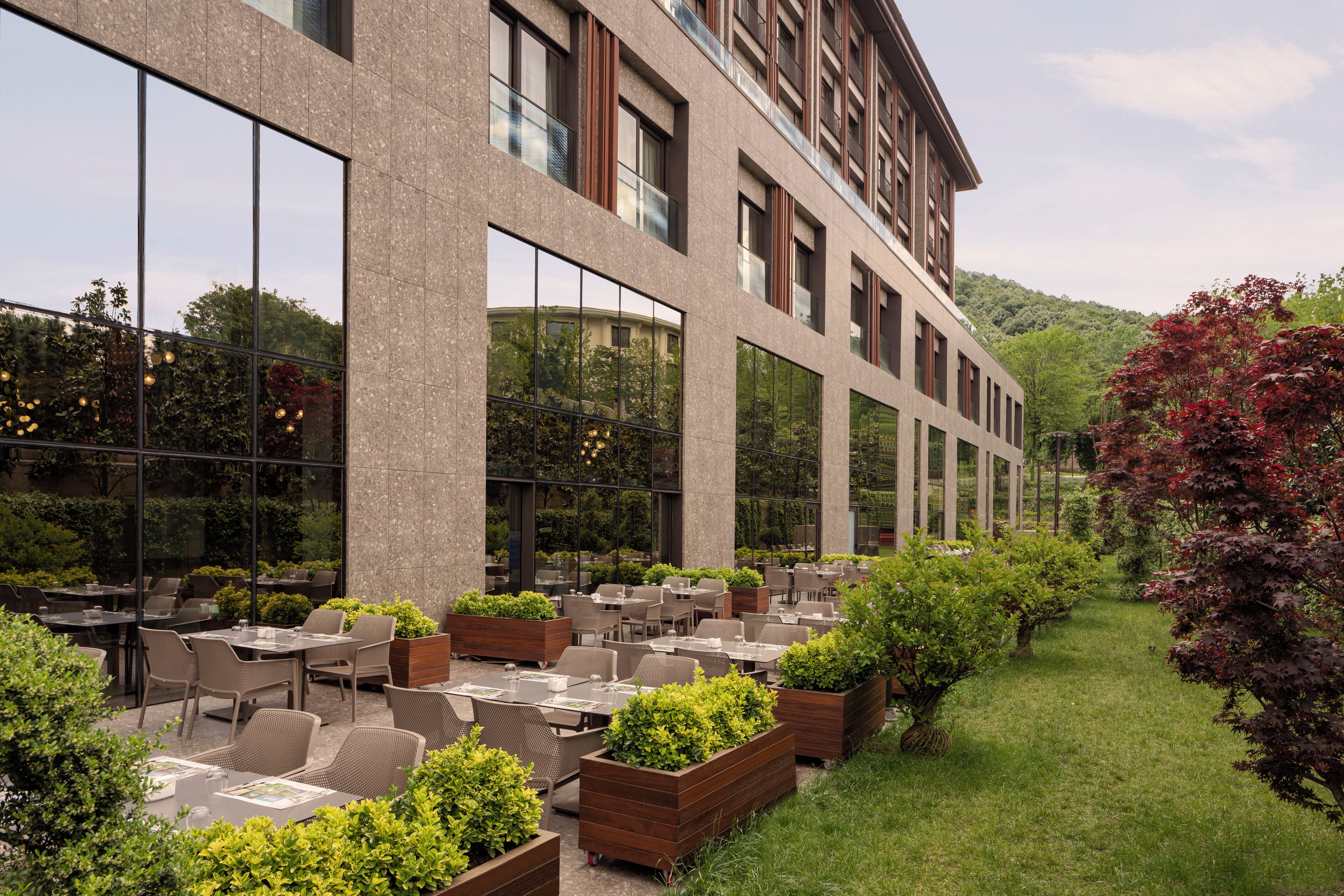 A modern outdoor dining area with tables and chairs, surrounded by greenery and a building with large windows.