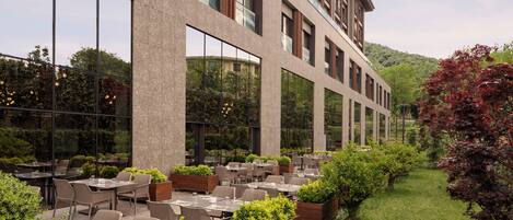 A modern outdoor dining area with tables and chairs, surrounded by greenery and a building with large windows.