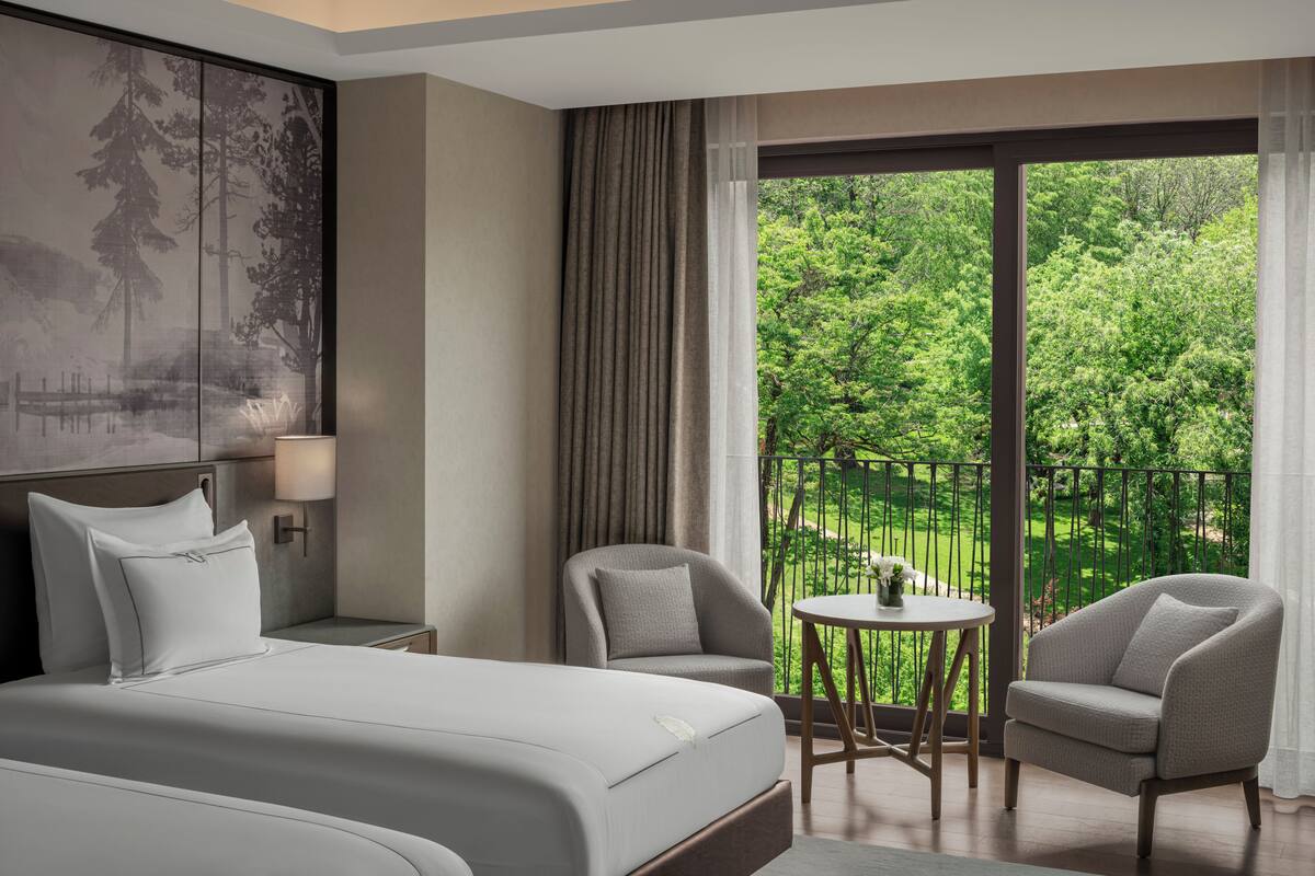 Ein Hotelzimmer mit einem großen Bett, zwei Sesseln, einem kleinen Tisch und einem Balkon mit Blick auf Grünflächen.
